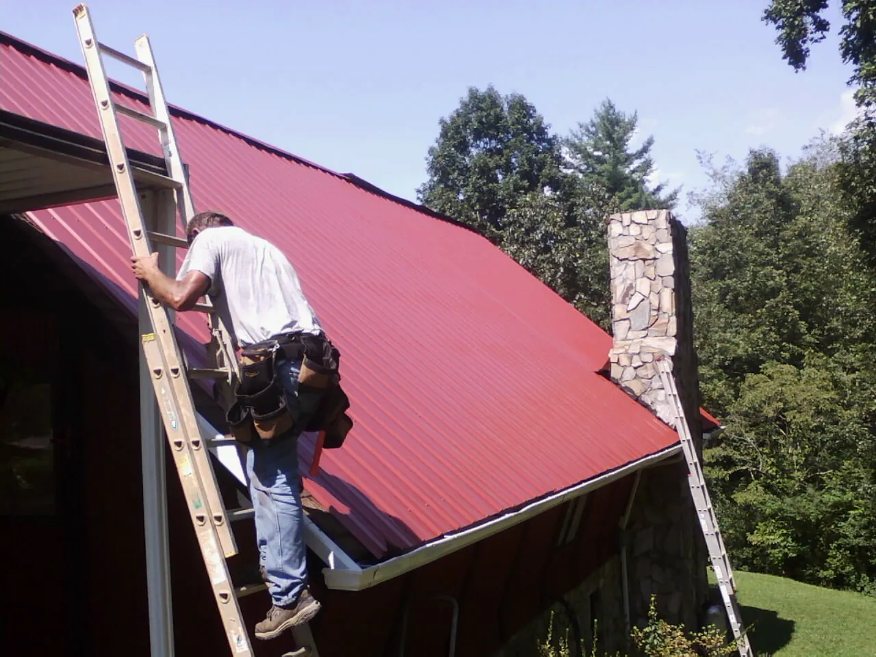 Roofer on a ladder beside a red metal roof and stone chimney — Soffit & Fascia Repair by Roofing Brothers in Sellers